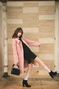 Fashion model in pink coat and black dress striking a dynamic pose indoors with stylish black heels.